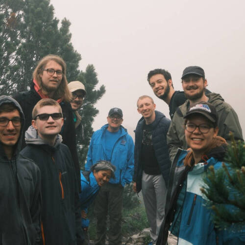 A group of eleven people dressed in outdoor and hiking gear pose together in a misty forest surrounded by pine trees. Most are smiling and appear to be enjoying the hike. The atmosphere is foggy, creating a serene and adventurous setting.